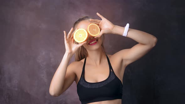 Funny Fitness Woman Posing with Half of Orange Covering Eyes Playing at Black Studio Background