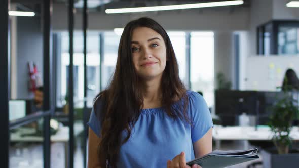 Portrait of caucasian businesswoman standing in office holding paperwork smiling to camera