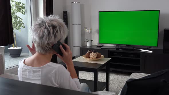 A Middle-aged Woman Sits, Watches TV with a Green Screen and Talks on a Smartphone