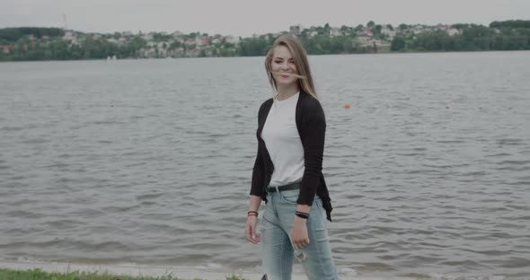 Cheerful Girl in Casual Clothes Walking at Riverside During Wind and Posing