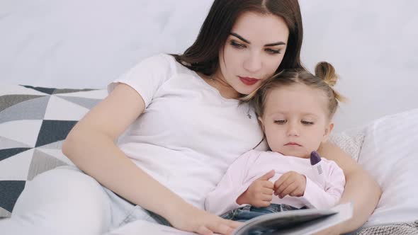 Beautiful Mother and Toddler Daughter Are in Bed Hugging and Reading a Book