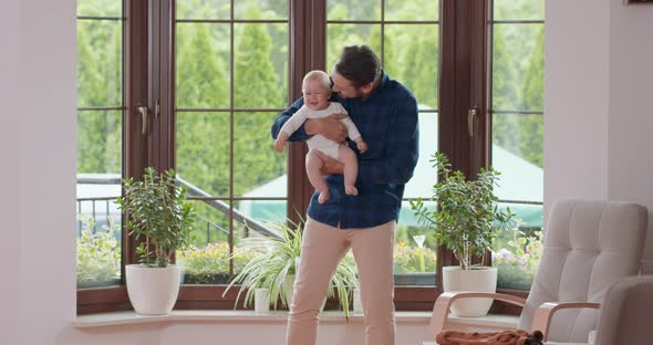 Bearded and Mustashed Father Holds His Adorable Baby and Slightly Waves on the Legs Smiling in Front