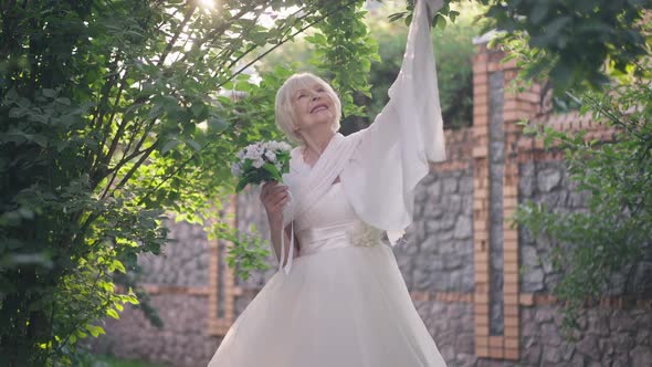 Smiling Happy Senior Bride with Grey Hair Standing in Sunshine Raising Hand with Shawl in Slow