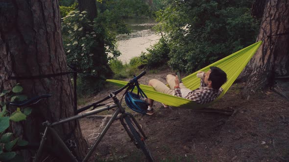 A Young Man Came to the Park on a Bicycle and is Resting in a Hammock Overlooking the Lake While