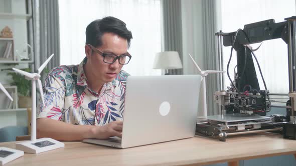 Asian Man With Laptop Computer Programming Electric Wind Turbine Model At Home