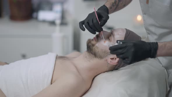 Side View Young Man Lying with Closed Eyes As Tattooed Male Beauty Therapist Applying Moisturizing