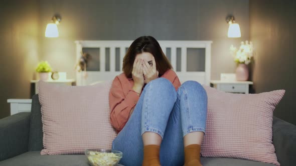 Scared Young Woman Sitting on the Sofa, Eating Popcorn, Watching TV and Covered Her Face with Hands