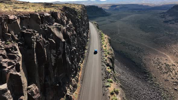 Wide drone shot of a bright blue car driving on a rocky highway.