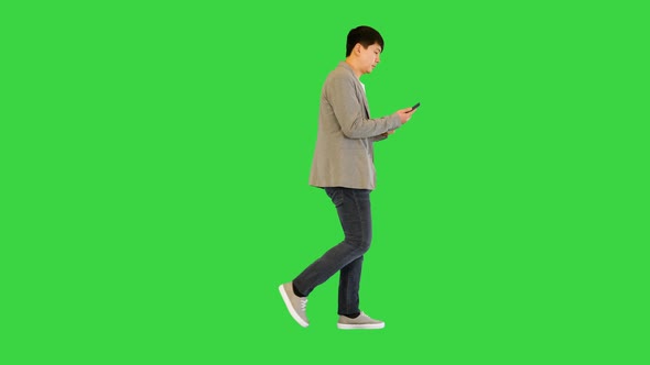 Young Asian Man Walks Looking at His Smartphone on a Green Screen Chroma Key