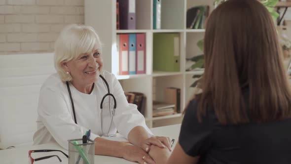 Elderly Woman Doctor Advises Young Woman Patient and Holds Her Hand in the Office