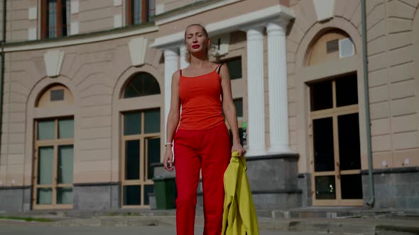 a Woman in Red Clothes Holds a Yellow Jacket in Hand and Walks Against the Background of a House