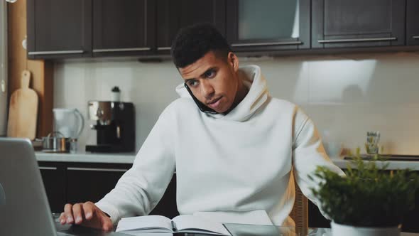 Mixed-race Man Speaking By Smartphone and Working on Computer at Home Kitchen