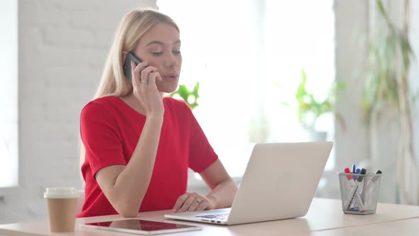 Young Blonde Woman Talking on Phone while using Laptop in Office