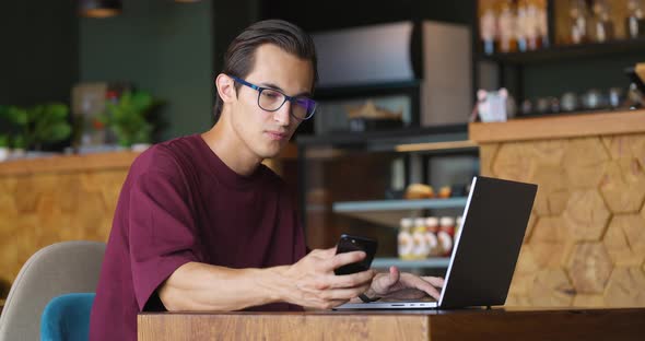 Young Man Working on Laptop and Checking Smartphone for Messages While Having Coffee Break in Small