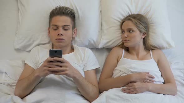 Angry Girl Looking at Boyfriend Using Cellphone in Bed, Internet Addiction