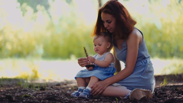 mother and baby sit in a park or in a forest on the ground looking at twigs and cones