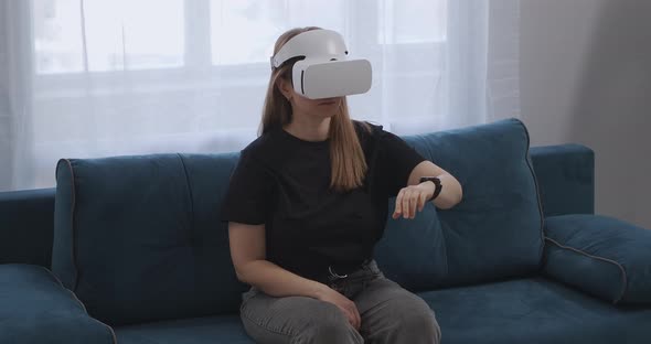 Woman with HMDdisplay on Head Is Sitting on Couch in Apartment Gesticulating By Hands Virtual