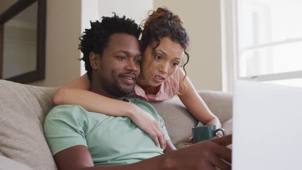 Worried biracial woman with coffee embracing her male partner sitting on sofa with laptop