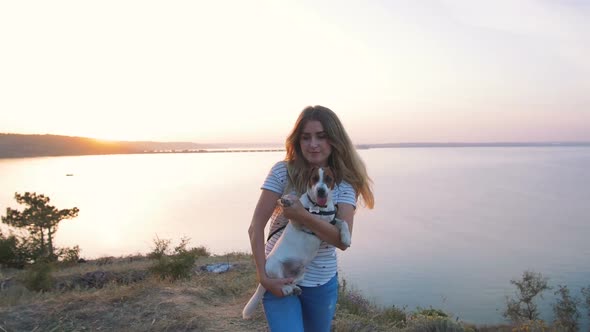 Young Attractive Woman Playing with a Dog Jack Russell in the Meadow at Sunset with Sea Background