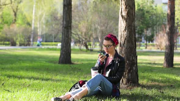 Beautiful Young Stylish Woman Sitting Under a Tree in the Park with a Cup of Coffee and a Telephone