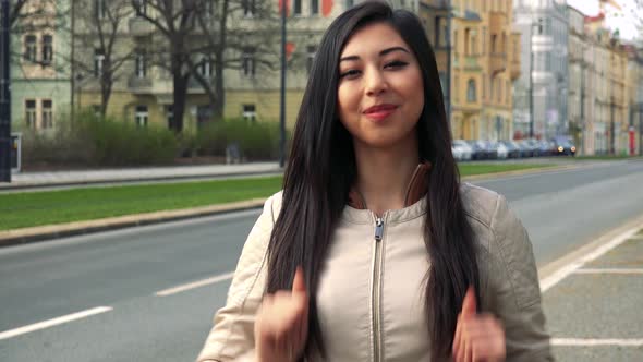 A Young Asian Woman Shows a Double Thumb Up To the Camera in a Street in an Urban Area