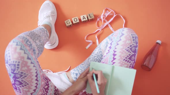 Motivating Word Is Laid Out On Orange Yoga Mat. Woman In Sportswear, Holds Notepad And Writes Down