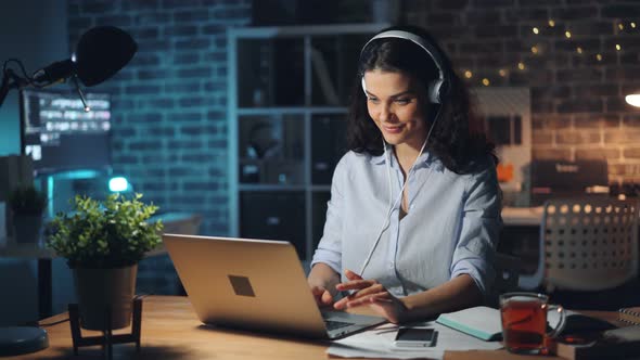 Pretty Lady in Headphones Listening To Music in Dark Office Working with Laptop