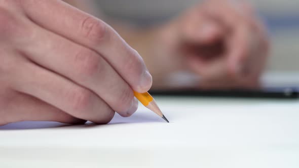 A student writing with a yellow pencil and surfing the internet on a mobile phone at a table