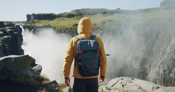 Rear View of Man in Yellow Jacket and Backpack Walking at Cliff Edge Looking at Detifoss Waterfall