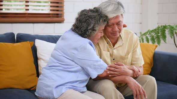 Asian senior elderly couple sitting on a sofa, woman giving hugging to man, support, understanding.