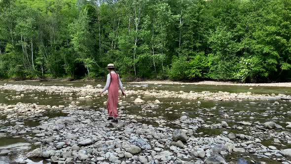 Vintage romantic woman in straw hat, long dress walks on river rocks near forest. Natural scenic