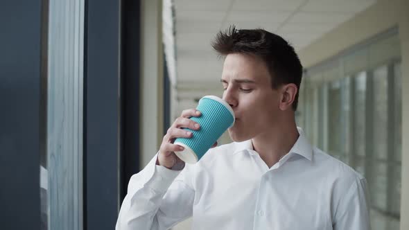 Happy Businessman Drinks a Cup of Coffee and Looks Out the Window in Office