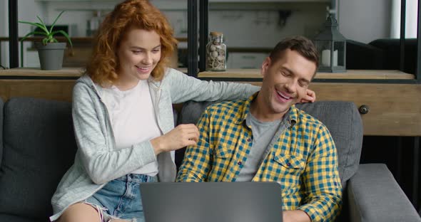 Young Woman Is Approaching and Sits Down Near Man with a Laptop on a Grey Couch