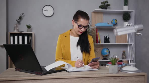 Businesswoman in Eyeglasses which Holding Phone in Her Hand while Working with Report with Graphs
