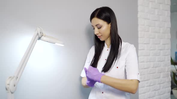 A cute cosmetologist in a white lab coat stands near the wall and disinfects gloves