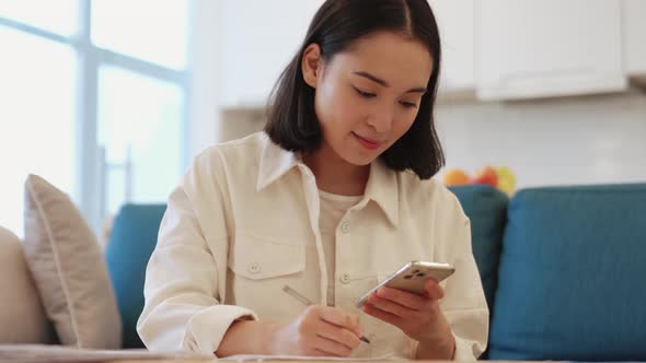 Cheerful Asian woman writing something from smartphone