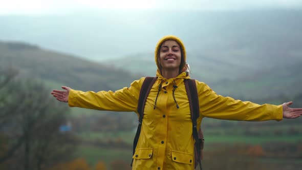 Beautiful Happy Smiling Woman in Yellow Raincoat Retro Backpack and Knitted Beanie Stands Outdoors