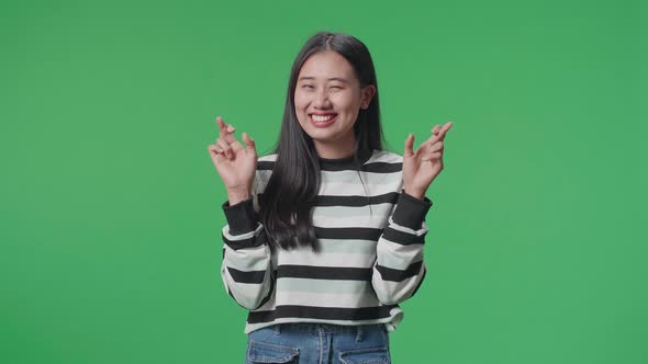 A Smiling Asian Woman Keep Fingers Crossed While Standing In Front Of Green Screen Background