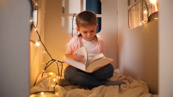 Cute Little Boy Sitting in Toy Cardboard House at Night and Reading Big Fairy Tale Book