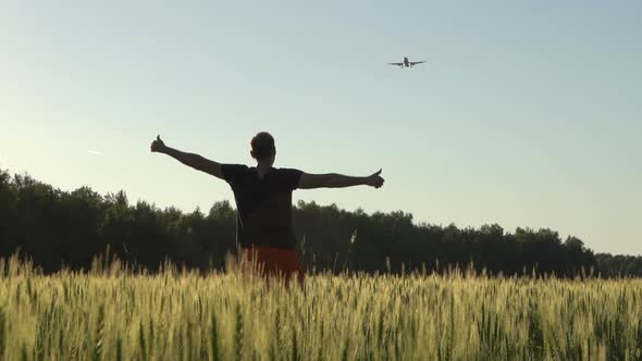 Happy Young Man Joyfully Greets an Airplane Flying in the Sky in a Wheat Field in the Summer at