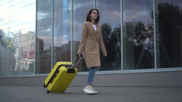 A Young Woman Walks with a Yellow Suitcase. A Girl with Glasses and a Coat Walks Along the Mirrored