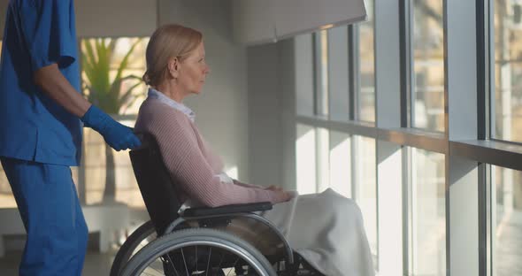 Nurse in Medical Mask and Gloves Pushing Wheelchair of Disabled Senior Patient at Hospital Corridor