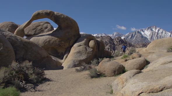 A young man trail running with his dog past a natural rock arch in a mountainous desert.