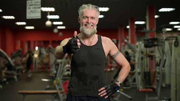 A smiling gray-haired respectable man with a beard and tattoos shows a thumbs up