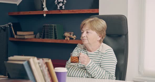 An Elderly Woman Looks Into a Computer Monitor While Working at Home and is About to Drink Tea