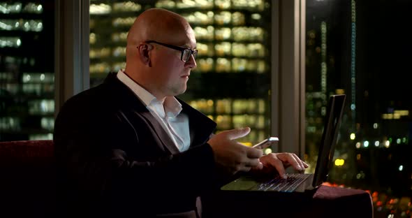 Bald Businessman Sits Near a Large Window. He Enters the Data From the Phone Into the Computer