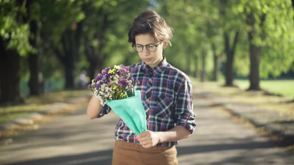 Portrait of Teenage Boy with Bouquet of Flowers Standing in Sunbeam in Summer Park Waiting for Date