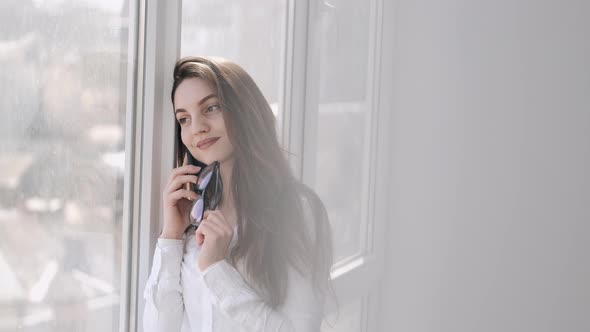 Pretty Woman in Glasses Is Making a Call Near the Window and Smiling