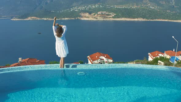 Beautiful Woman in White Dress Stands By the Pool Edge and Enjoys the Summer View To the