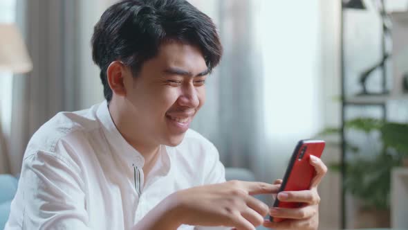 Close Up Side View Of Asian Man Touching Smartphone And Laughing While Sitting On Sofa